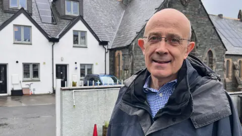 Peter Toes, chairman of Keswick Community Housing Trust. He is stood outside a development the trust built for locals. He is bold and has blue eyes and wears glasses. He is wearing a blue and white chequered shirt and a blue coat. 