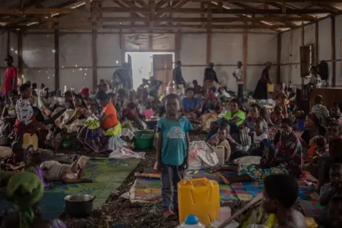 JOSPIN MWISHA / AFP People stand crowded into a darkened makeshift home. They have very few belongings. Many are children.