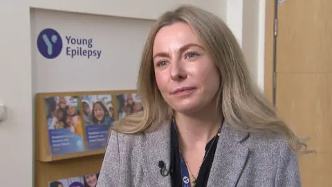 Mark Norman BBC Lara Carr wearing a grey jacket and black blouse with Young Epilepsy brochures on a shelf in the background 