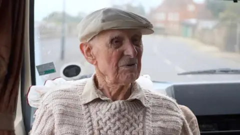Bill wearing a cream cap and a beige jumper and shirt sat inside a van with the road behind him