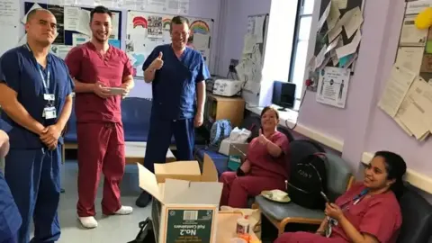 Peter Dutch Five hospital workers congregated around an open cardboard box. Two are wearing blue scrubs, and three are wearing mauve scrubs.