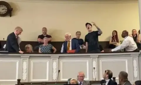 A video still of members of the public sat in a gallery above councillors in the city council chamber. One man, dressed in a black T-shirt and a black hat, is stood up and gesturing with a raised arm towards the councillors.