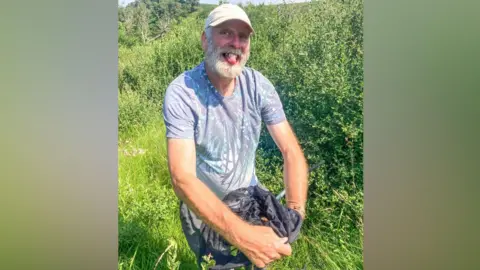Charlotte Rankin Dave Wainwright looking up at the camera and smiling while holding the small moth within a black net. He is wearing a cream cap and has a white beard. He is wearing a faded blue t-shirt with what looks like a butterfly design on it. He is standing in a field with long grass behind him.