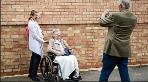 Getty Images Woman in wheelchair and carer having their photo taken in front of the memorial wall at Bletchley Park