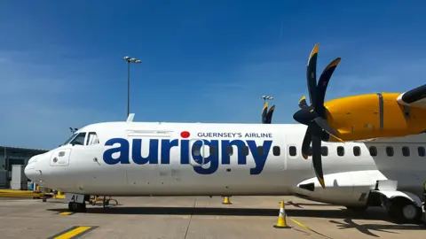 A white turboprop aircraft with yellow tail and engine nacelles, parked on an airport apron under a clear blue sky. The fuselage displays the word “aurigny” in large blue letters with a red dot above the “i,” and the text “Guernsey’s Airline” in smaller font. The aircraft has two black propellers, and a ground crew member wearing a high-visibility vest is standing nearby. Traffic cones are placed around the plane.