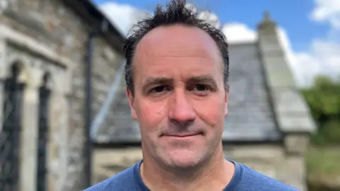 Man with short black hair looking at the camera with the church behind him 