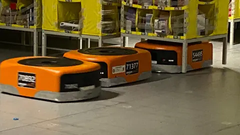 Orange robots, the size of small boxes, run along a warehouse floor, under yellow shelving stacks, which they run under, attached to, and move around. 