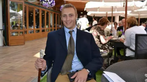 Contributed A man with short blonde hair sits in a chair at a table and smiles. He wears a navy suit jacket with a blue shirt and gold and dark tie. He holds a staff in one hand with the top of it carved into the shape of a duck's head.