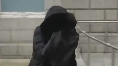 A person wearing a parka walks outside a court building covering her face in a black material