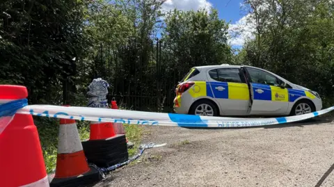 Stuart Woodward/BBC Blue and white police tape put in place across a road with a parked police car behind the cordon. There are orange traffic cones to the left of the frame. There are also trees behind a metal gate or fence.
