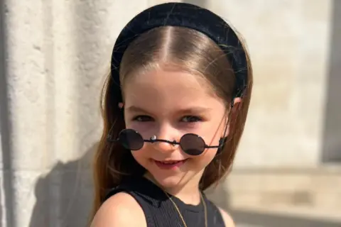 Ruka Family Leonna looks at the camera, with black sunglasses resting at the end of her nose. She has brown hair and brown eyes, and is standing outside in front of a stone pillar.