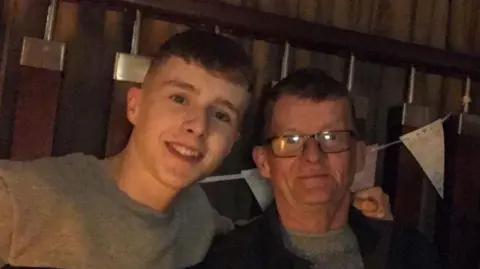 Matthews McGrath has short brown hair, his smiling and wearing a grey sweatshirt with his arm over his father. Tom McGrath has short brown hair, he is wearing black square frame glasses, a grey jumper and a black jacket. Behind them is white bunting tied to a set of bannisters. 