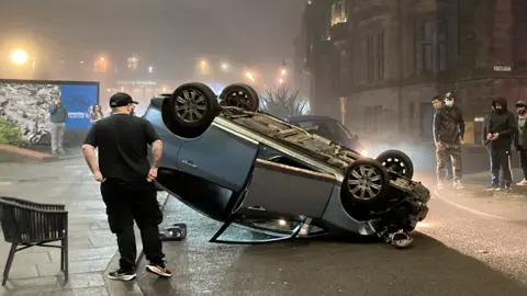 A silver car on its roof on a narrow street with people standing nearby, some on their phones