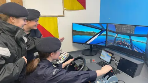 Three female sea cadets using a ship simulator. There are two screens in the photo with a steering wheel and controls. 