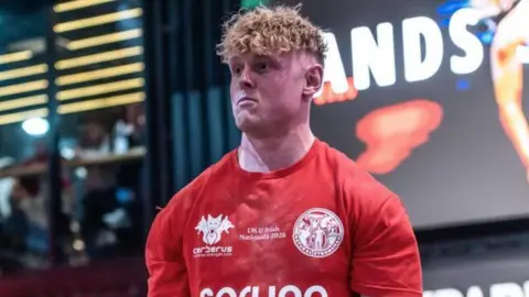 Jake Orr, a man with blonde curly hair, is pictured in a red sports top. 