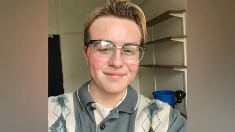 A selfie of a young man wearing glasses in a small university accommodation bedroom. He is wearing a patterned jumper and is smiling at the camera.