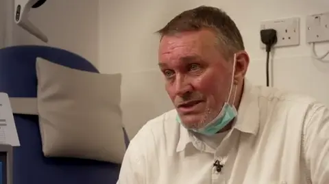 A man sits on a hospital bed in a white short. He is receiving treatment at the hospital and equipment can be seen in the background. 