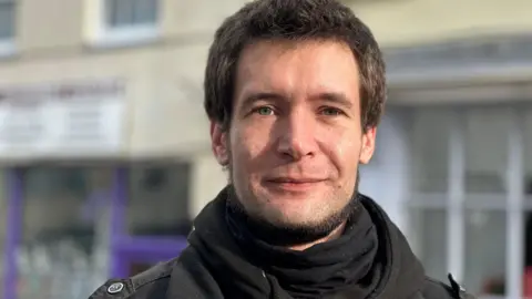 Man with dark hair wearing a black coat and scarf 