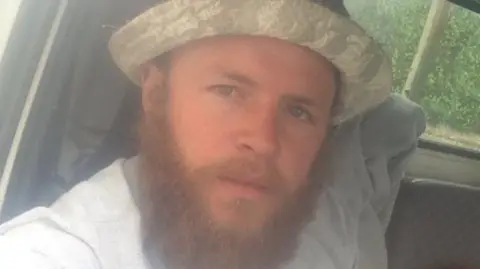 Facebook Kirk Bishop - a man with a full, reddish-brown beard and moustache, wearing a grey T-shirt and light camouflage brimmed hat. He is looking directly at the camera with his blue eyes.
