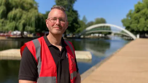 Alex Pope/BBC Bjorn Hove, standing by a river, with short hair, glasses and has on a black T-shirt and red high-vis jacket. He is smiling. A large path is to the right and a bridge in the distance. 