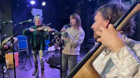 Musicians from the Bristol Ensemble practice at the Trinity Centre. One woman is playing a violin, another is playing the clarinet and in the foreground a man in a cream jumper is playing a cello
