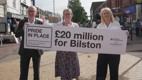 Pat McFadden MP, Josie Kelly and councillor Linda Leach stand in a street in Bilston town centre holding a board or banner saying £20m for Bilston, Pride In Place. 