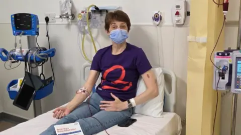 A woman with short dark hair and wearing a face mask sits on a hospital bed in blue T-shirt and jeans, hooked up to a red tube connected to a transfusion machine.