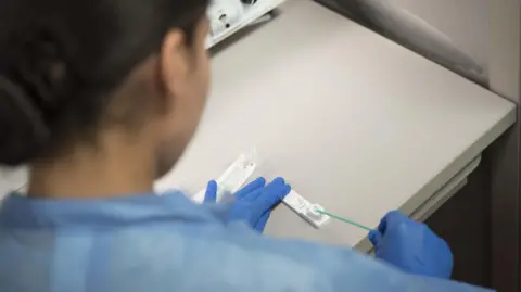 A picture taken over a woman's shoulder. She appears to be wearing medical clothing, including blue plastic gloves. She is inserting a long swap into a white unit on a desk. 