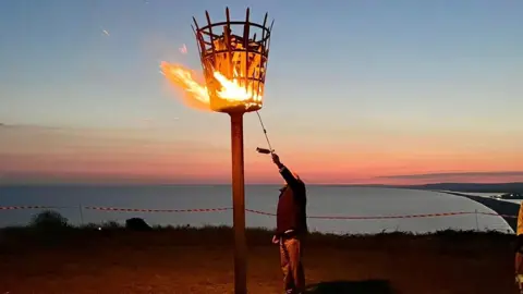 A man reaching up to light a beacon - a basket on a pole which is already burning well. The beacon is on top of a hill and backdrop is the coast and Chesil Beach. It is dusk and the sky is pink and orange.