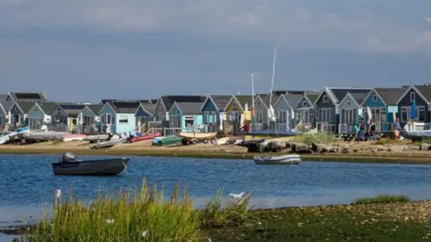 Getty Images View across Christchurch Harbour towards Mudeford Sandbank which is packed with large beach huts with small boats laying on the sloping beach.