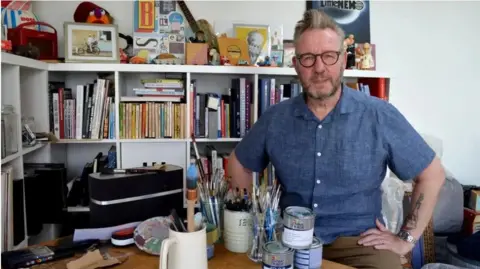 BBC Artist Pete McKee in his studio in Sheffield wearing a blue shirt and brown trousers, surrounded by paints and brushes