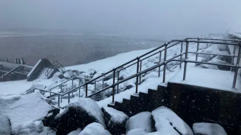 BBC/Jessica Lane A snowy scene of the promenade with steps going down onto the beach which is covered in a couple of inches of snow.