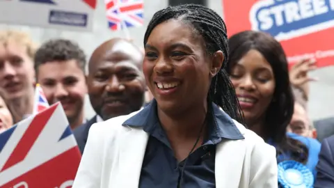 A grinning Kemi Badenoch celebrates a victory in Westminster with activists waving union jack banners and wearing blue Tory rosettes