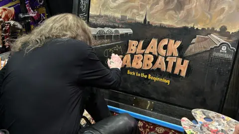 Luke McDonnell Ozzy Osbourne is leaning forward to sign his name on the Black Sabbath painting.