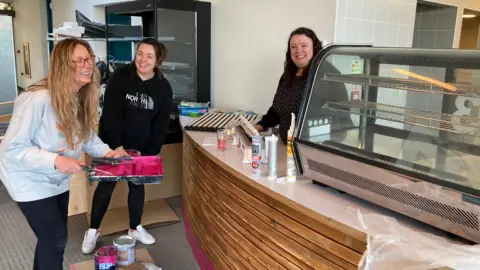 Three women smile at each other. One is behind a café counter, another is bent over, holding a paint tray and roller.