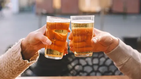 A stock photo of two hands both holding pints of lager as they cheers their glasses.