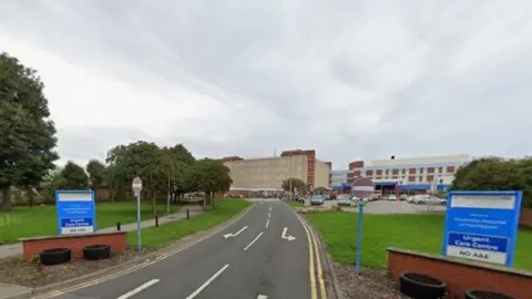 A road leading to the entrance of Hartlepool Hospital. There are two blue signs and on one of them it says there is no A&E. The hospital s in the background and is spread across two or three buildings.