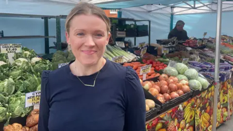 A woman with tied back blonde hair and a dark blue top in front of a stall with lots of fruit and vegetables on it