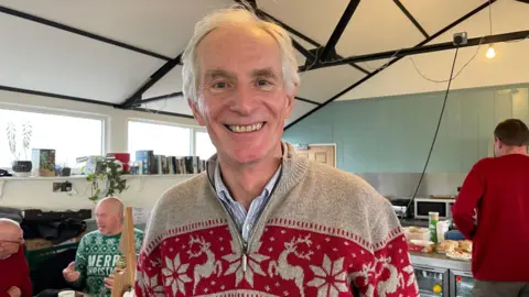 Martin Thomas has grey hair and is smiling. He is wearing a grey and red Christmas jumper with a reindeer and snowflake pattern on it.