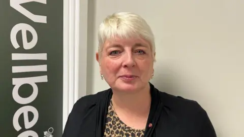 Ollie Conopo/BBC Sheralee Webb with short blonde hair and gold earrings wearing a gold and black dress and black cardigan, standing beside a sign that says "Wellbeing".