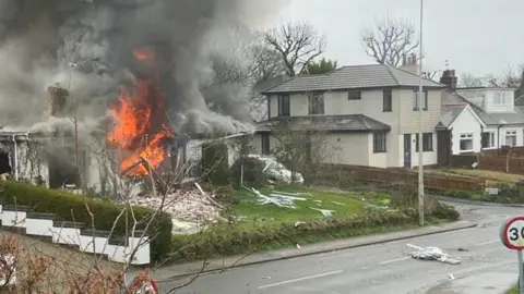 Martin Gardner House in flames on a street - billowing grey smoke rises from the single-storey white building