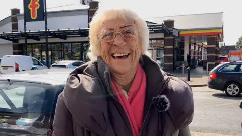 George King/ BBC A close-up head and shoulders image of resident Pat, who is laughing. She is standing outside a supermarket and wearing a purple coat with a pink scarf.