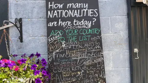 A chalkboard in the farmyard at Castle Farm asks "How many nationalities are here today?" Underneath in multicoloured chalk is written Peru, North Korea, New Zealand, Czech Republic, Bulgaria, Bangladesh and Sweden amongst many others.