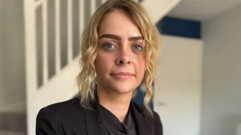 A woman with blonde curly hair, wearing a black jacket and shirt. She is standing in a house, in front of a stair case.