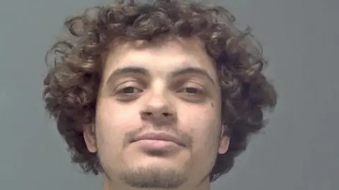 Suffolk Police Logan Burnett smiles at the camera with his head tilted upwards. He has dark, curly hair and brown eyes. He has a small amount of facial hair. 