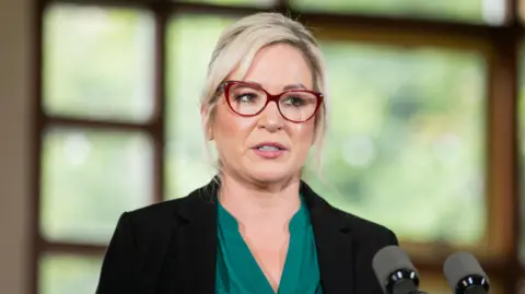 PA Michelle O'Neill in black blazer and green blouse with red glasses