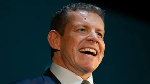 Matthew Horwood/Plaid Cymru Rhun ap Iorwerth speaking. He has a blue jacket over a white shirt. He is smiling in front of a dark background.