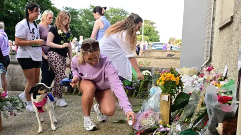 Pacemaker A woman placing a candle on the ground beside a pile of flowers. There are five women behind her and a dog beside her. 