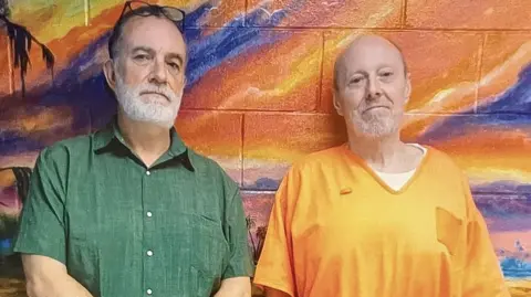 Rhys Wililams (l) - a white man with dark hair and a white beard and moustache stands alongside a balding man, also with a white beard and moustache, wearing an orange prison-issue top. The pair lean against a wall which has been painted and shows an orange, pink and purple sky.