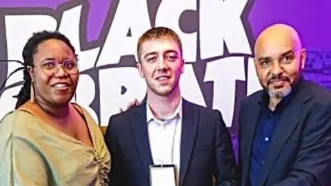 Birmingham City University A woman and two men standing in front of a large purple sign. The woman has dark hair and a beige top, the man in the middle has short brown hair, a white shirt and blue jacket and the man on the right has a bald head and beard and a blue suit